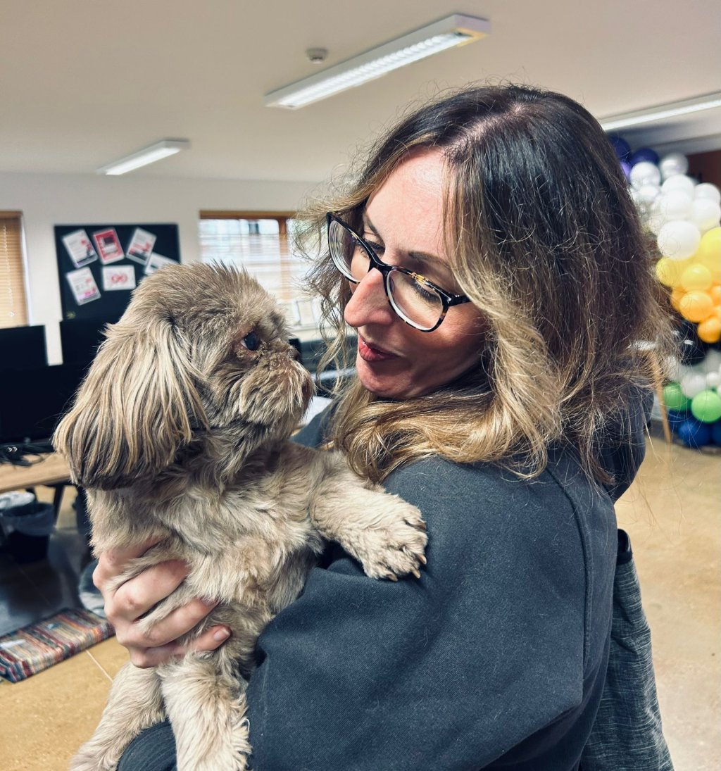 An HR consultant is holding a dog in her arms. She is standing in the middle of an office space.