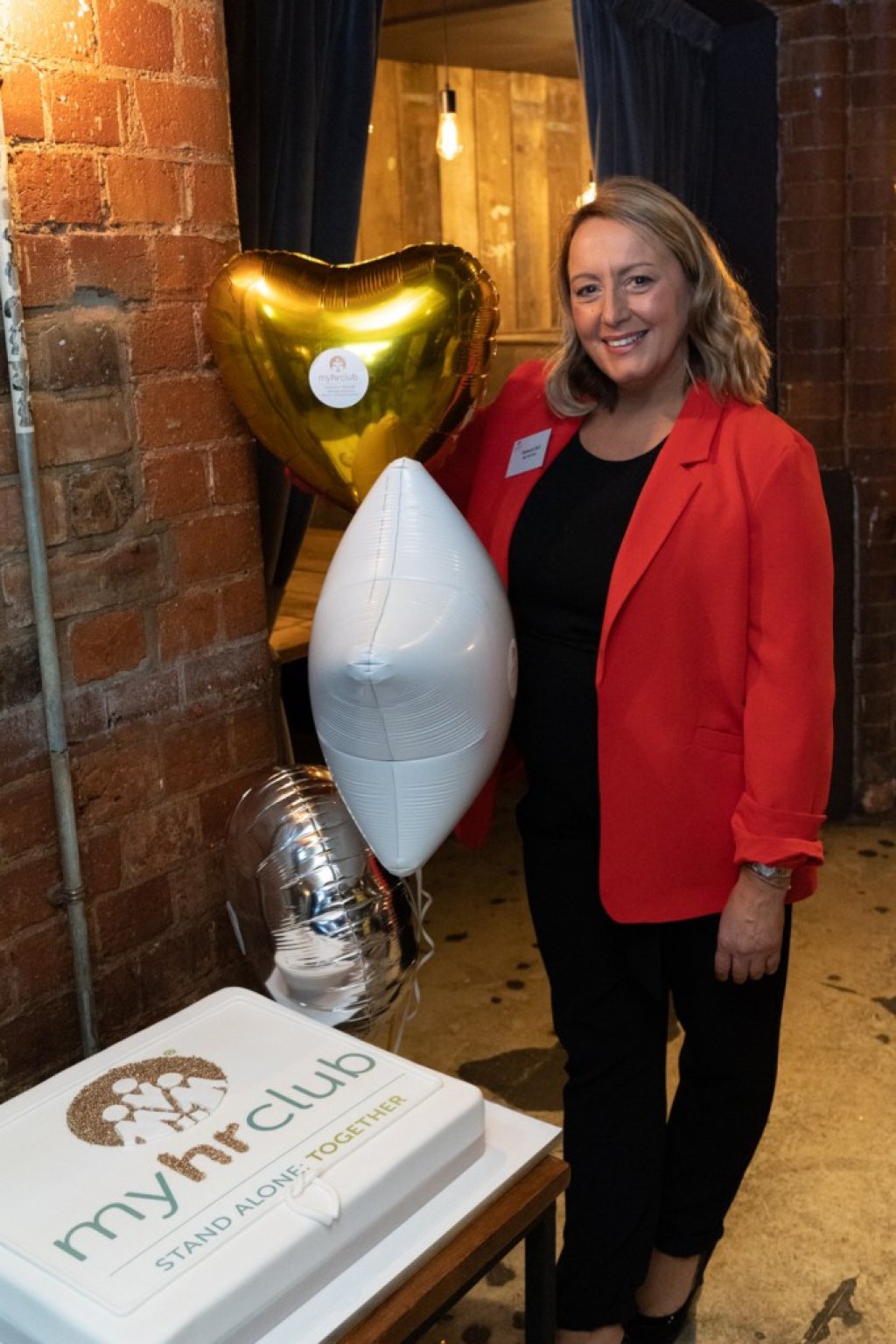 Founder of My HR Hub, Rebecca Bull, is standing next to balloons and a cake in celebration of the My HR Club launch.