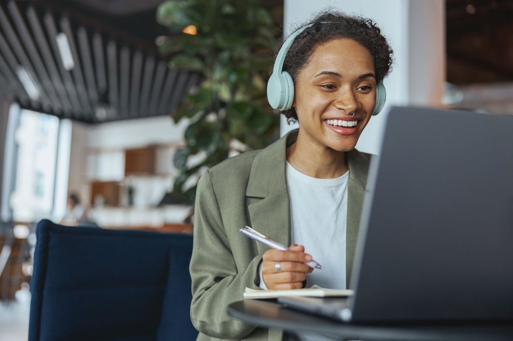 A woman is on a Zoom call on her laptop in a modern office space. She is smiling and is wearing overhead headphones.