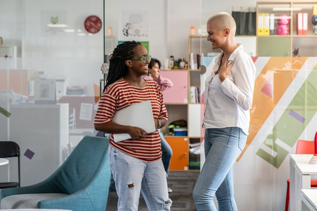 Two women are standing, talking in an office space. One is holding a laptop and is wearing large framed glasses. The other is in a casual shirt and jeans.