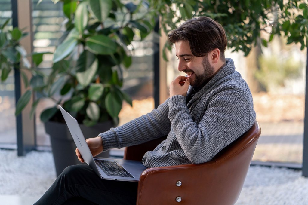 A man is sitting in a leather chair in a modern office space and has his laptop open on his lap.