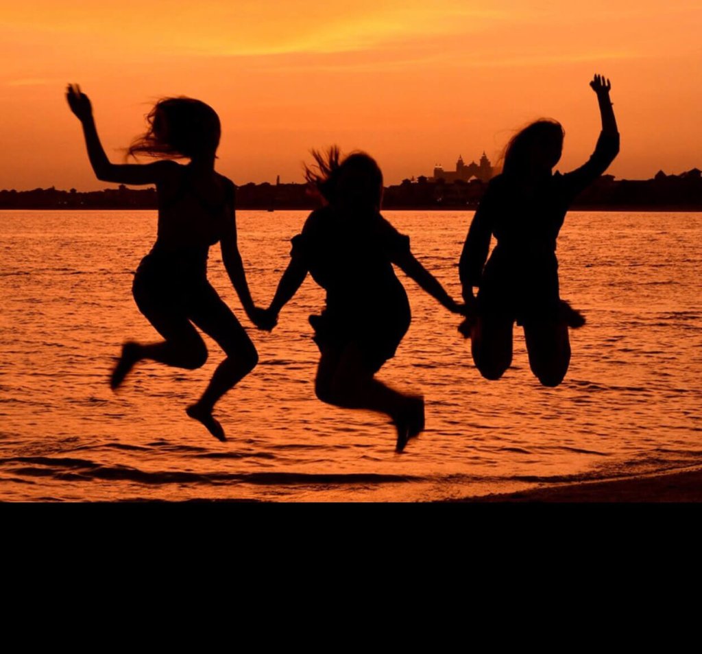 3 women are jumping hand in hand on a beach at sunset.