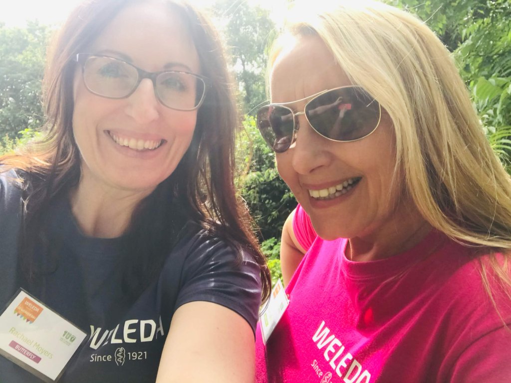 2 women are smiling as they stand side by side outside. They are both in Weleda t-shirts and sunglasses.