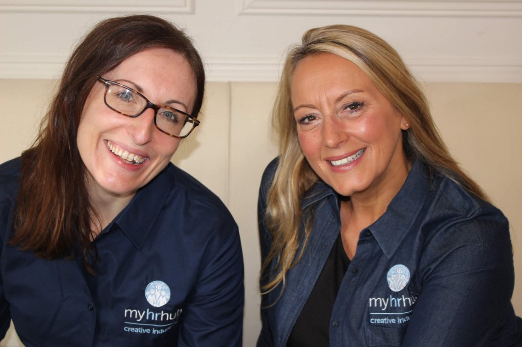 Two female HR consultants are sitting next to each other on a sofa and are smiling.